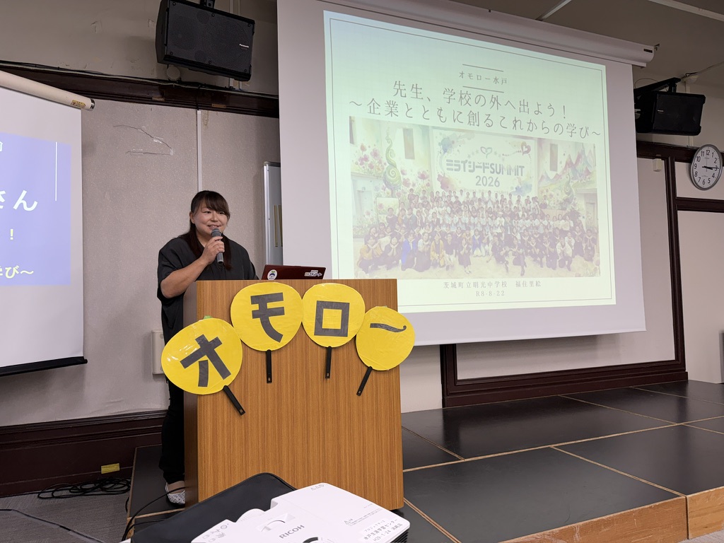 オモローと書かれた黄色いうちわがある演台でマイクを持ち先生、学校の外へ出ようという題名のスライドを背に発表する福住里絵さん