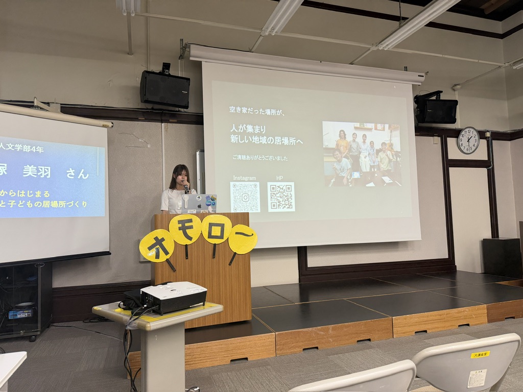 講堂のステージで発表する大学生の高塚美羽さんと空き家を地域の居場所に変える取り組みが映し出されたスクリーン