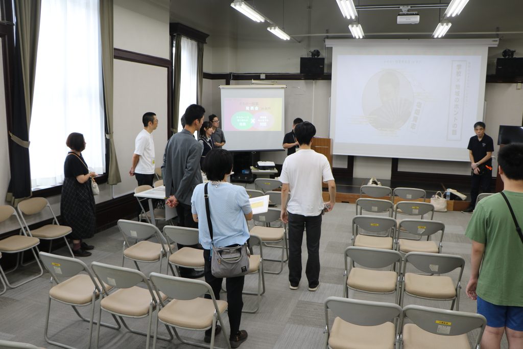 オモロー授業発表会in水戸の会場。学校と地域のホントの話という文字が映るスクリーンの前で人々が並び立っている様子