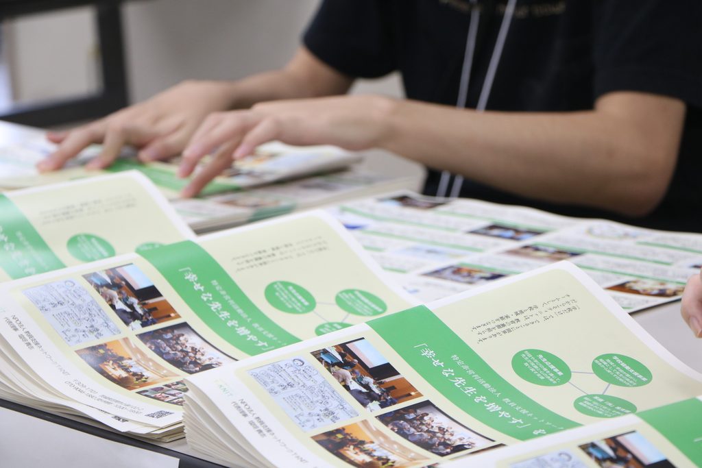 幸せな先生を増やすという言葉が書かれた緑色のパンフレットを机に並べて整理する様子