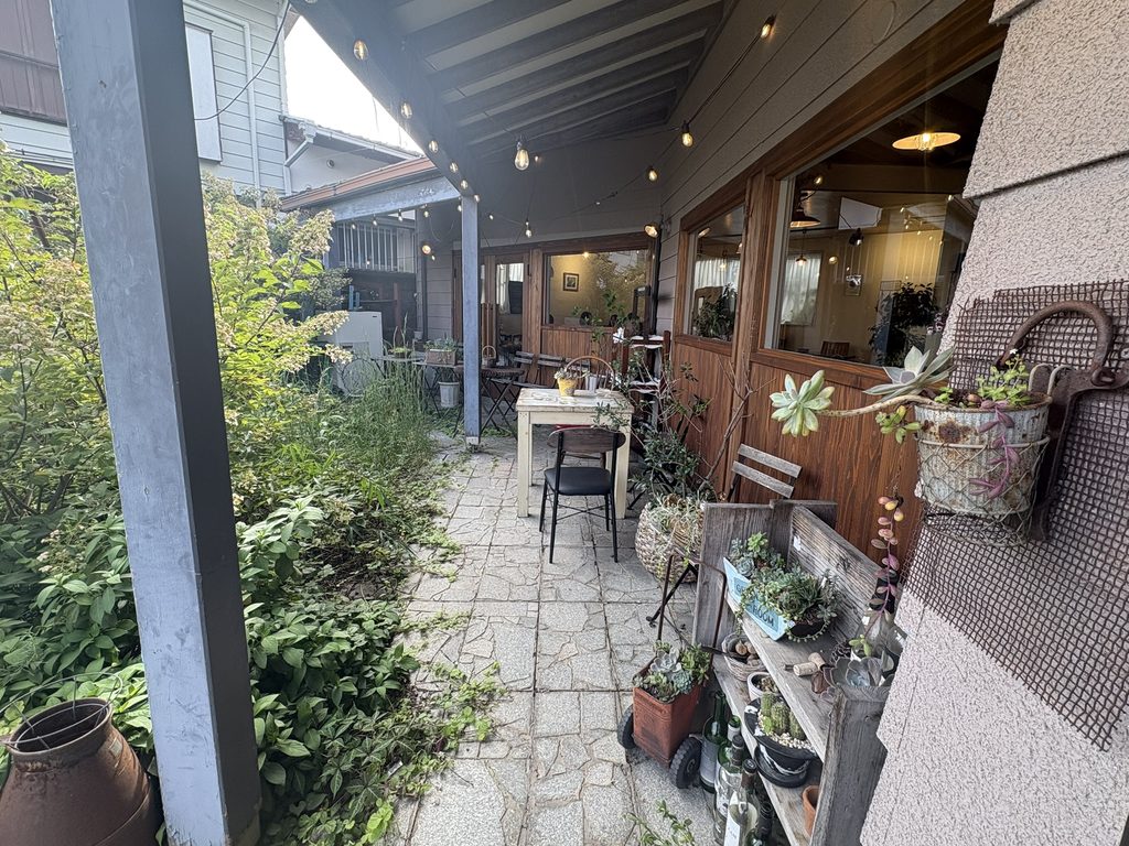 軒下にある屋外の座席と白いテーブルの周囲に多肉植物や緑の草木が並んでいる様子