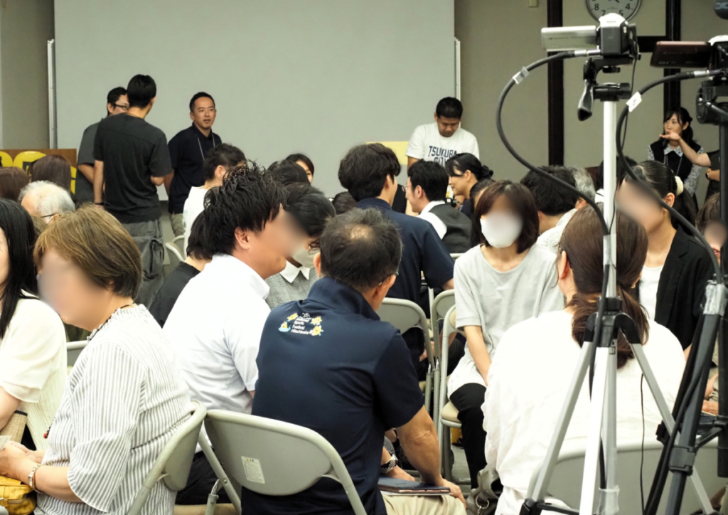 多くの人が集まる室内でパイプ椅子に座る人々と三脚に固定されたビデオカメラ