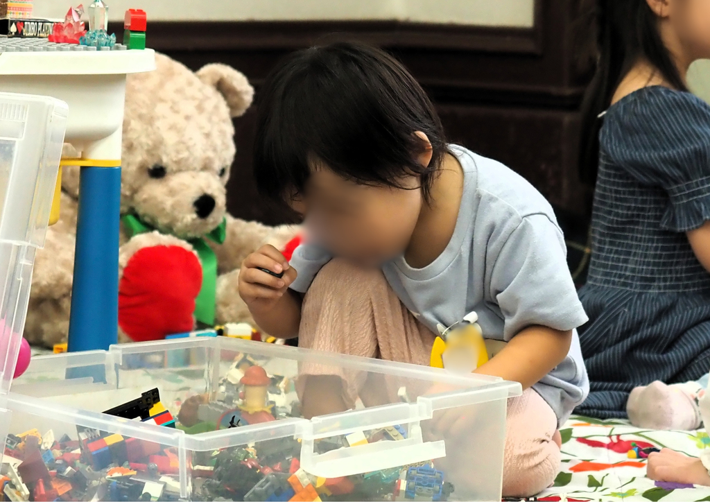 透明な箱に入ったブロックのおもちゃを手に取り遊ぶ子供と後ろに置かれた大きなぬいぐるみの様子(オモロー授業発表会in水戸のキッズスペース)