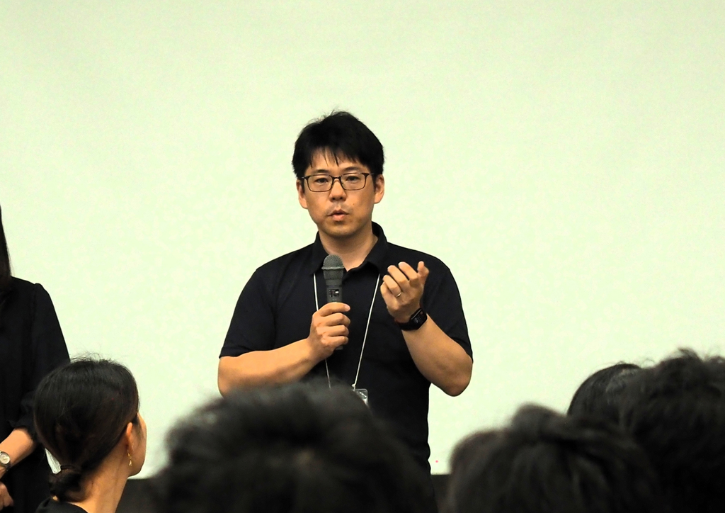 黒いシャツを着て眼鏡をかけた白土瑞樹さんがマイクを持って人々の前で話している場面